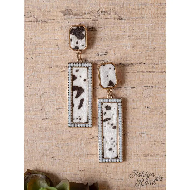 Pair of earrings with rectangular design and cow print pattern on a textured beige background.