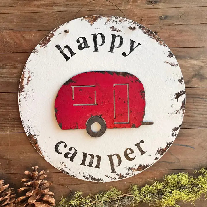 Round metal sign with red camper illustration and 'happy camper' text on a wooden surface.