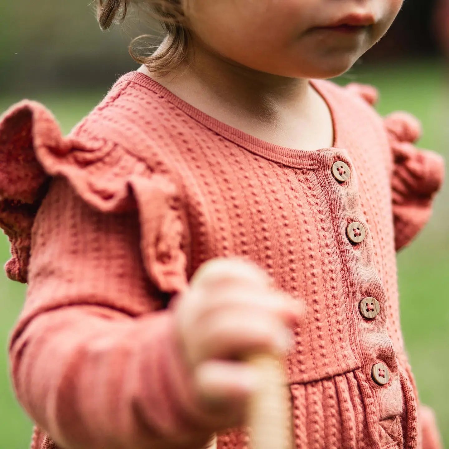 Child wearing a pink knitted sweater holding a wooden object outdoors