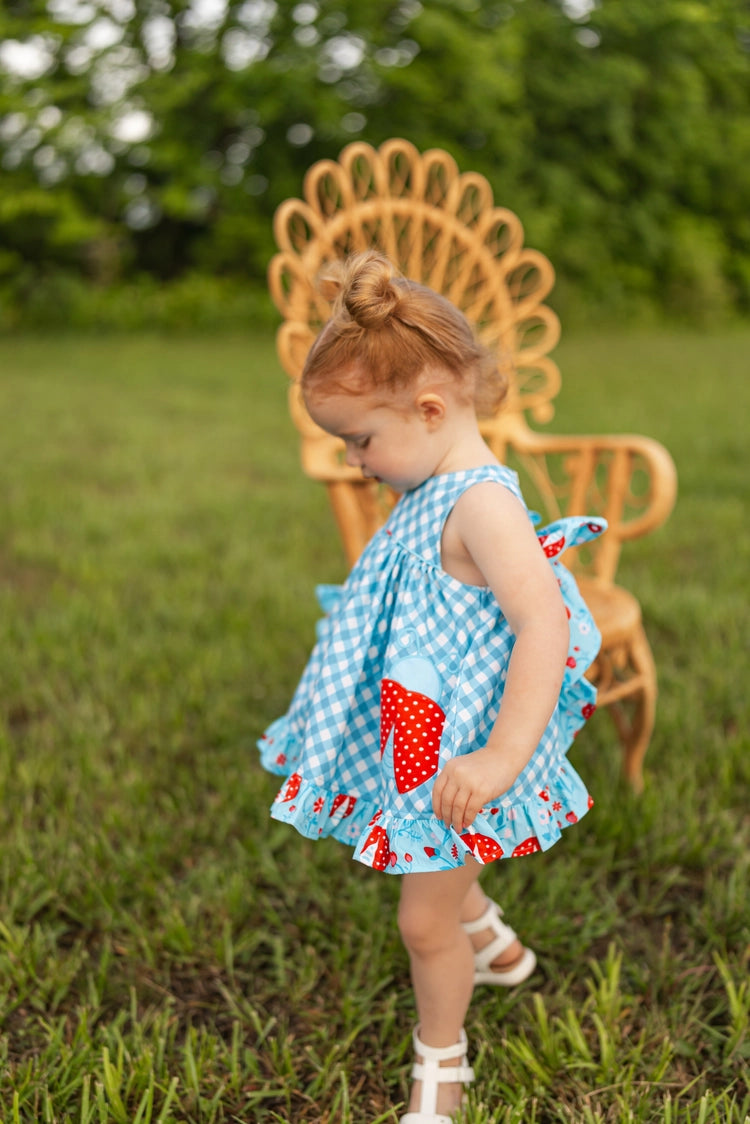 Child in a blue dress with red polka dots standing in a grassy field with a wooden chair in the background.