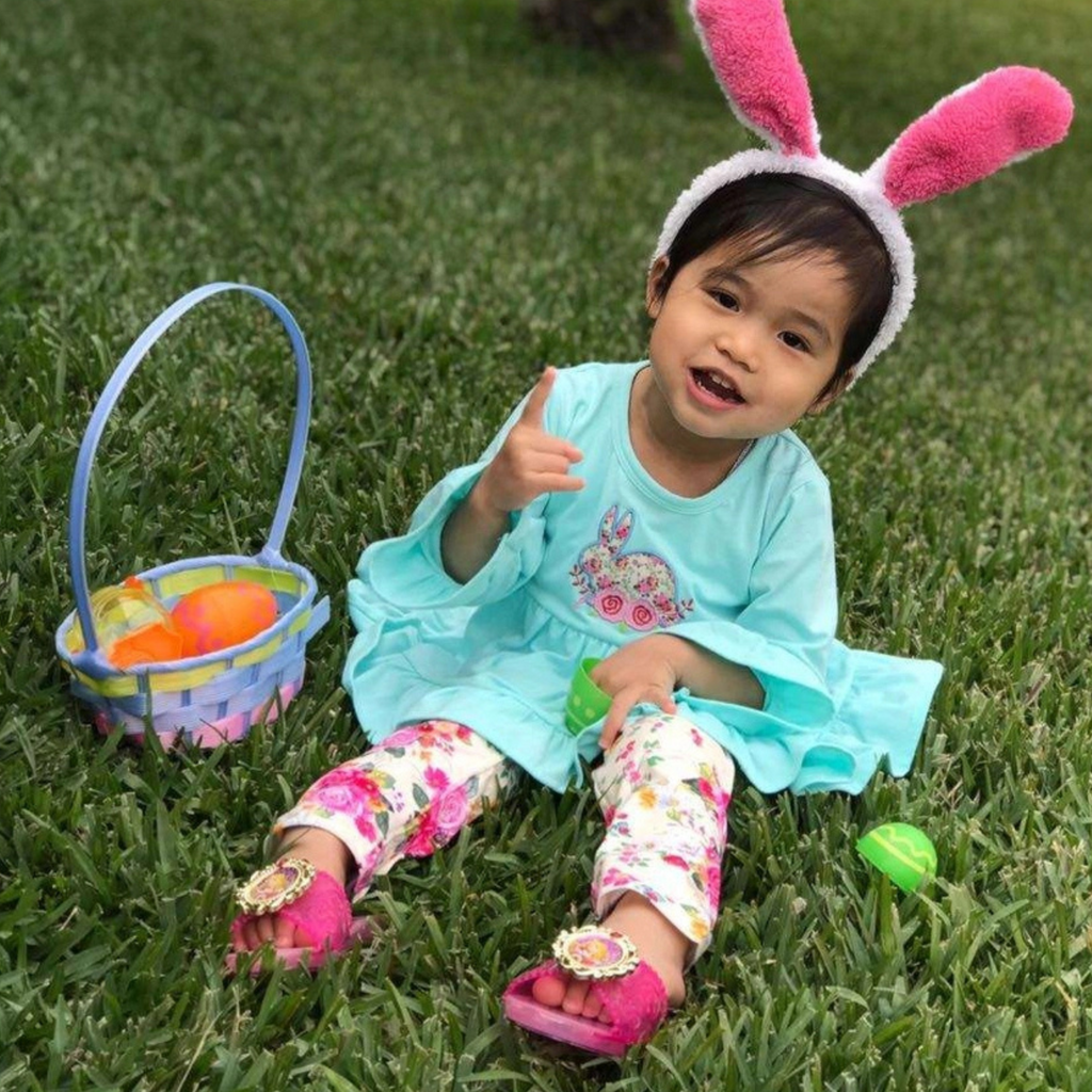 Child wearing bunny ears and a dress, sitting on grass with Easter eggs and basket.
