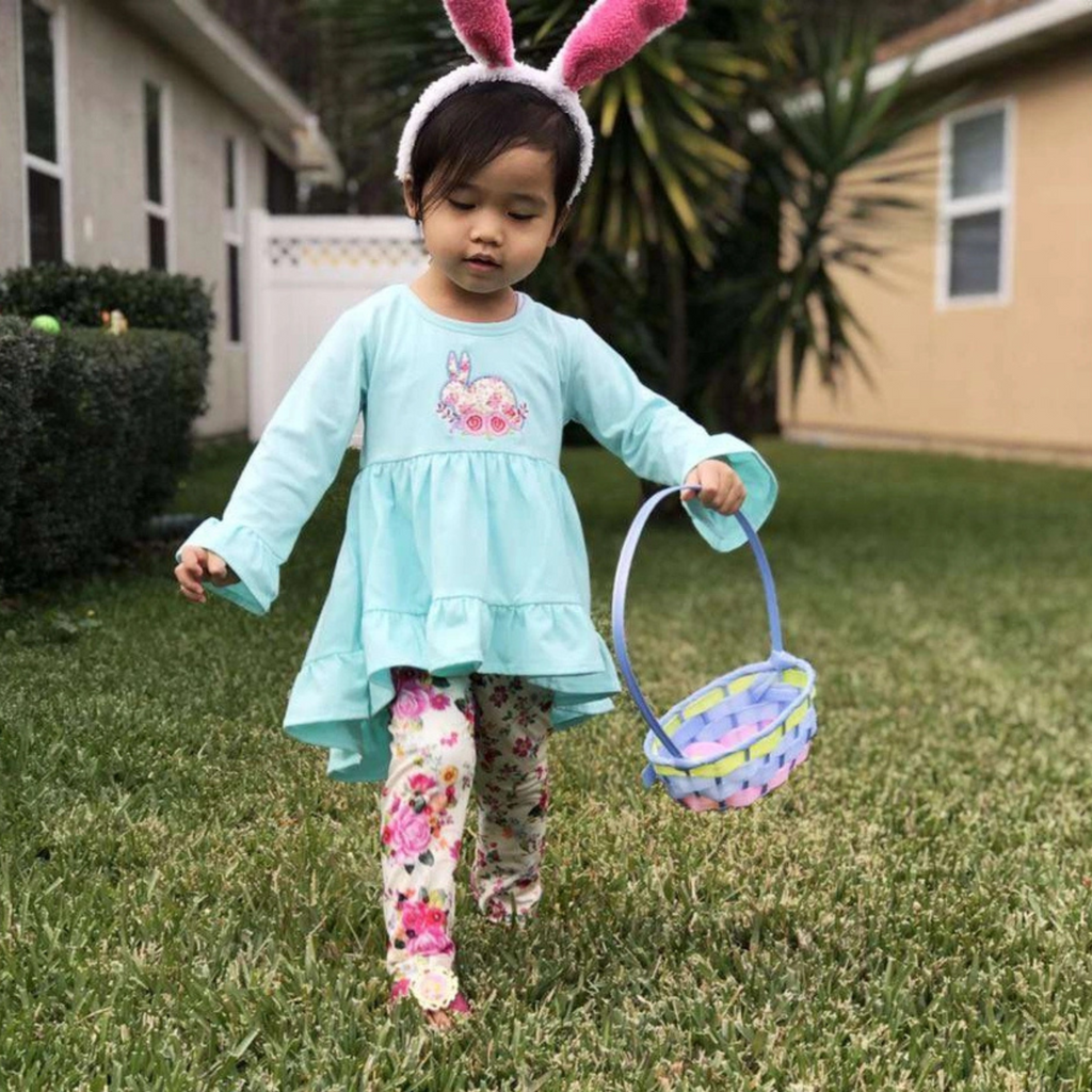 Child wearing bunny ears and a light blue dress with a floral pattern, holding an Easter basket outdoors.