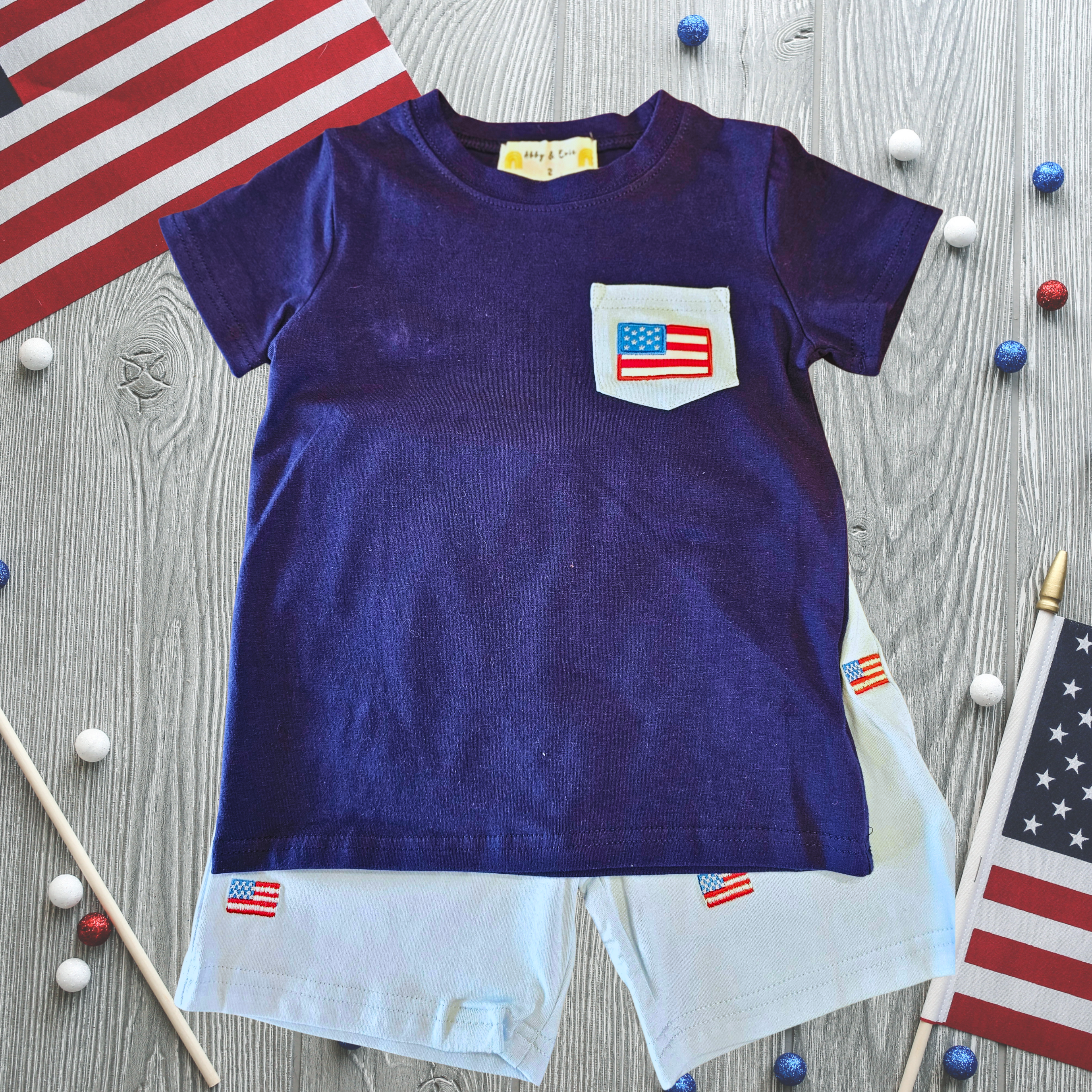 Children's blue t-shirt with American flag pocket and shorts on a wooden surface with flags.