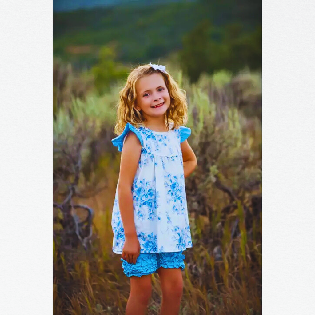 Girl wearing a blue floral top and blue shorts in a forest background.