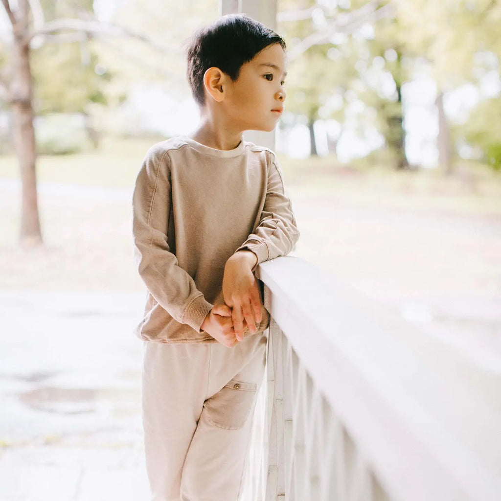 Child in beige outfit standing by a railing with a blurred natural background