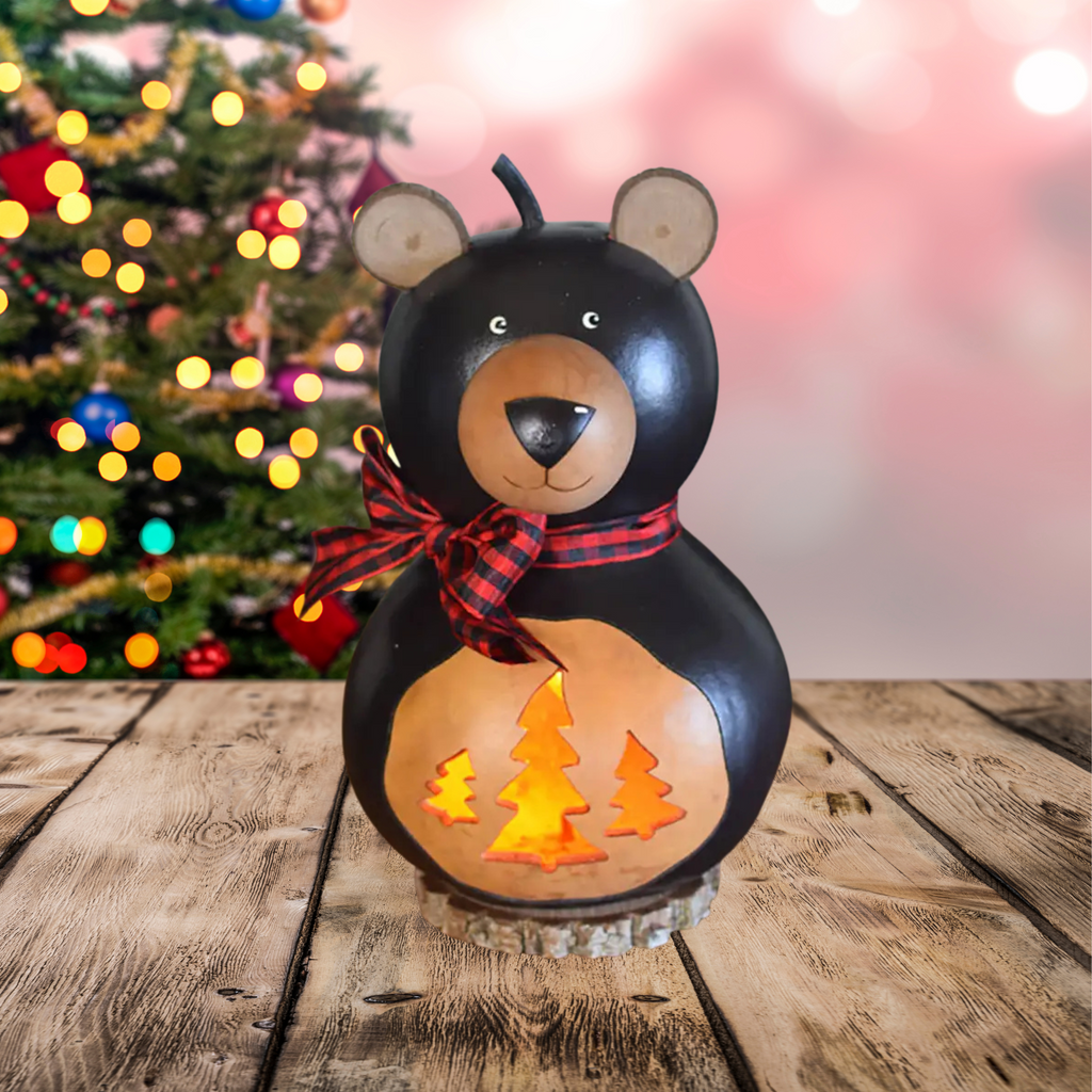 Decorative small bear-shaped gourd lantern with a bow, Christmas tree design, and blurred Christmas tree in the background.