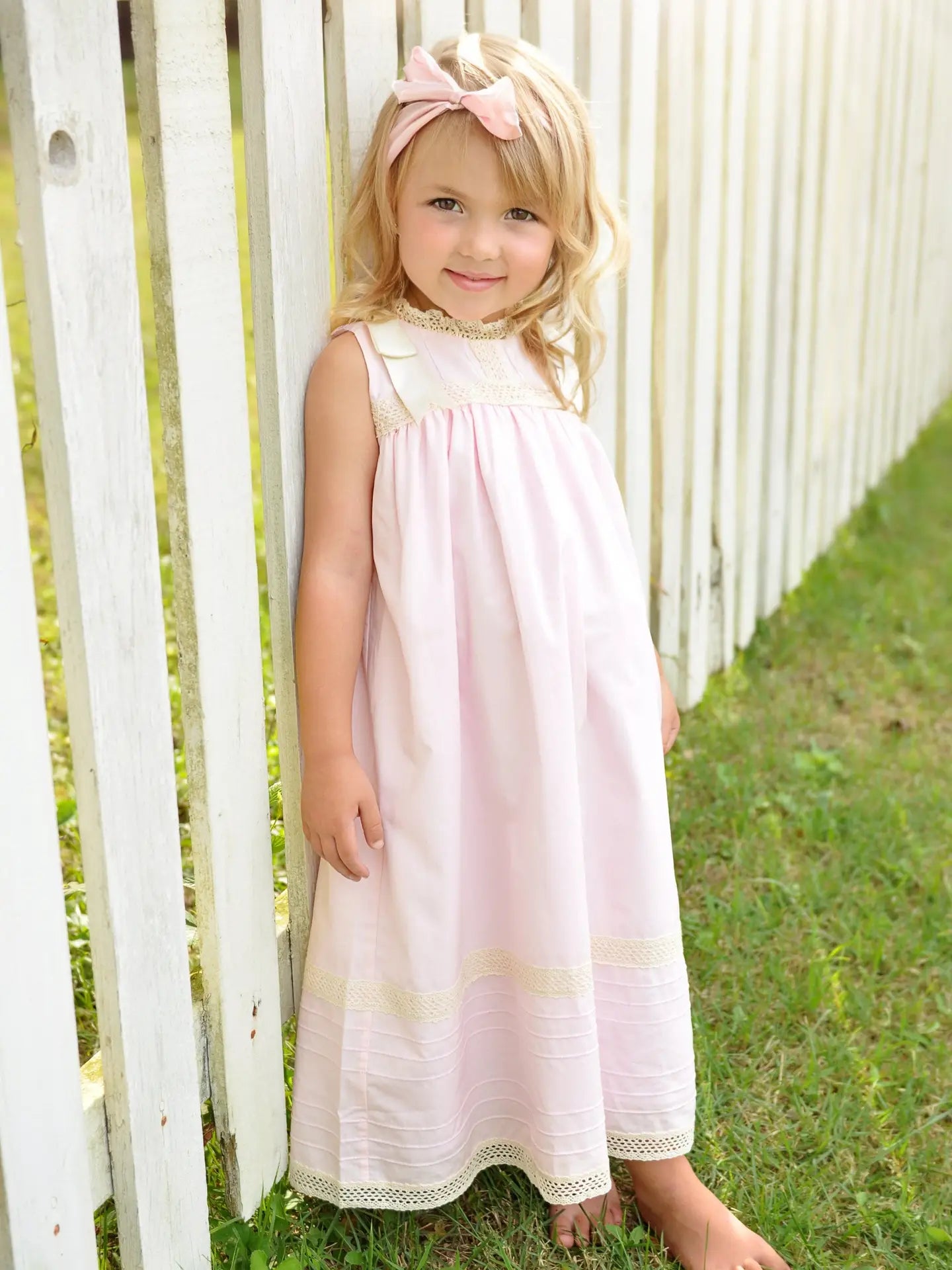 Young girl in a pink dress standing in front of a white picket fence.