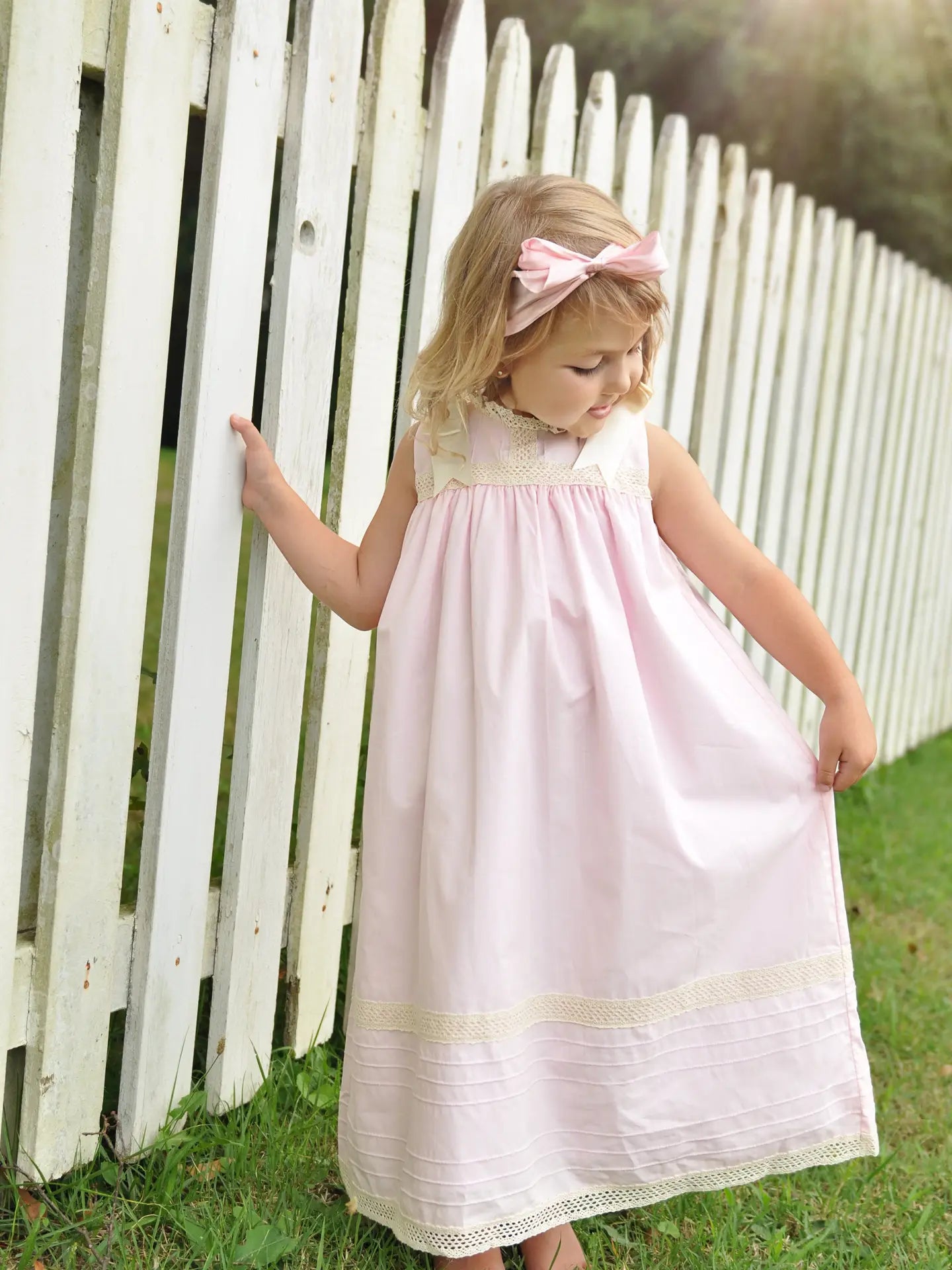 Young girl in a pink dress standing in front of a white picket fence.