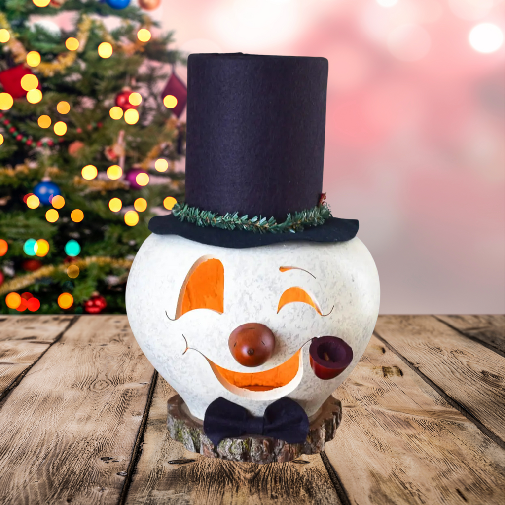 Decorative snowman with a black top hat with wreath, an orange carrot nose, red pipe, black bowtie on a wooden surface and a plug showing with a blurred Christmas tree in the background.