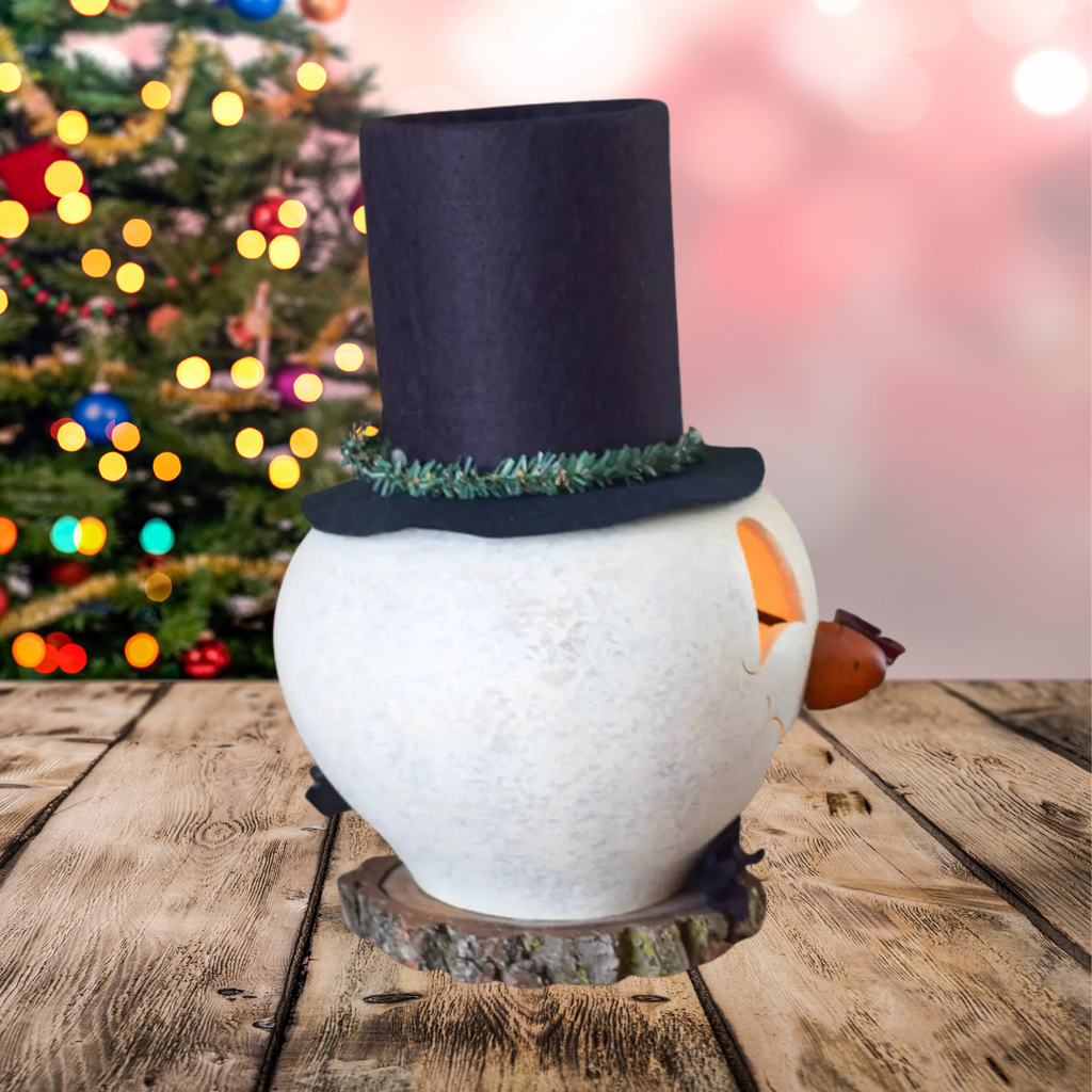 Decorative right side profile snowman with a black top hat with wreath, an orange carrot nose, red pipe on a wooden surface and a plug showing with a blurred Christmas tree in the background.
