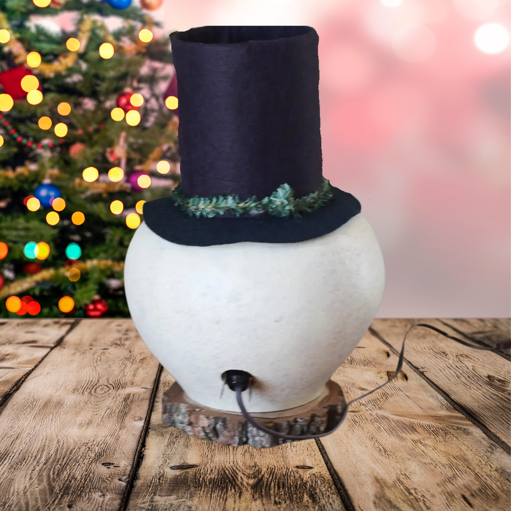 Decorative snowman with a black top hat on a wooden surface and a plug showing with a blurred Christmas tree in the background.