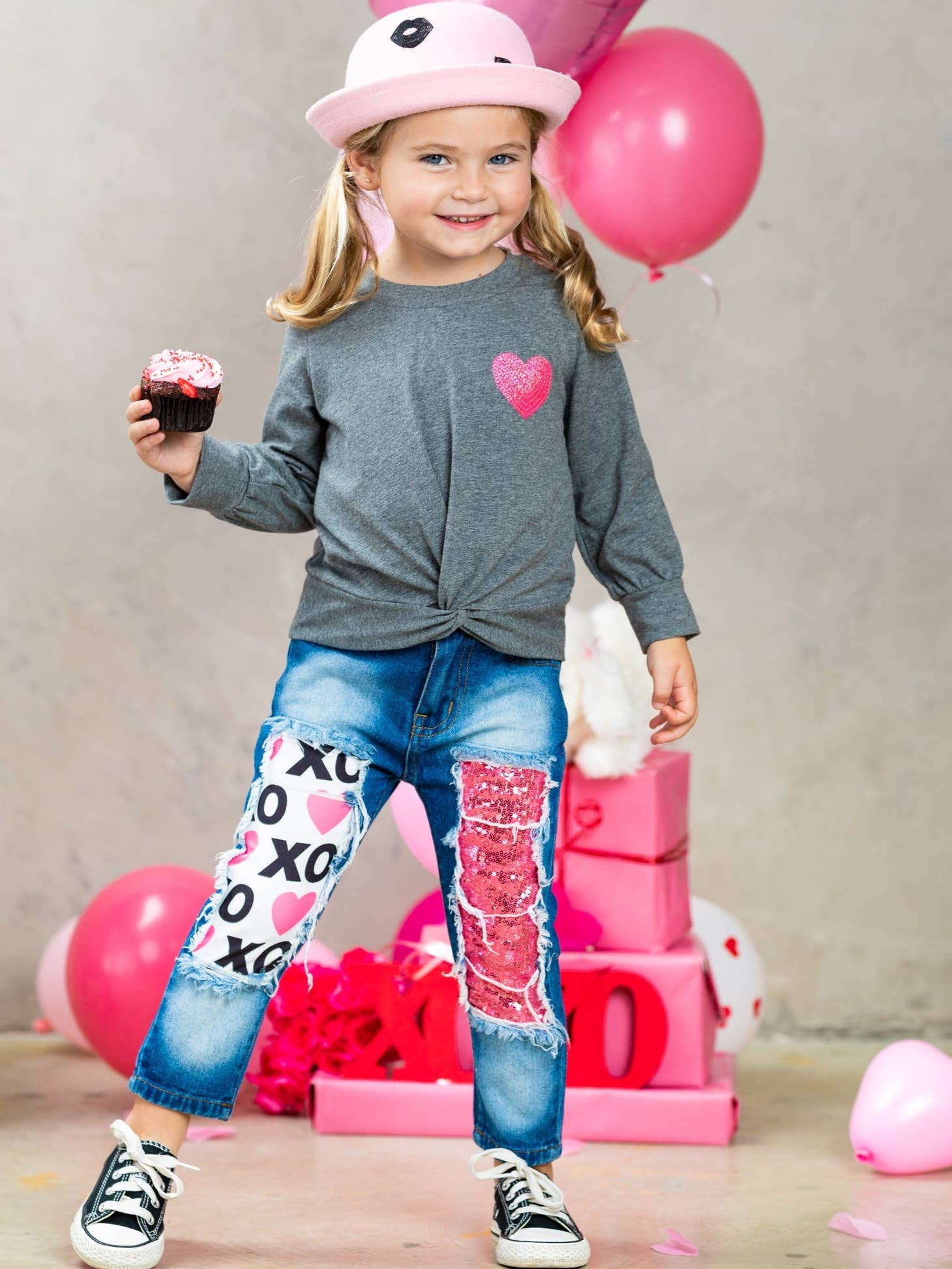 Child wearing a gray shirt with a heart design and blue jeans with heart patterns, holding a cupcake.