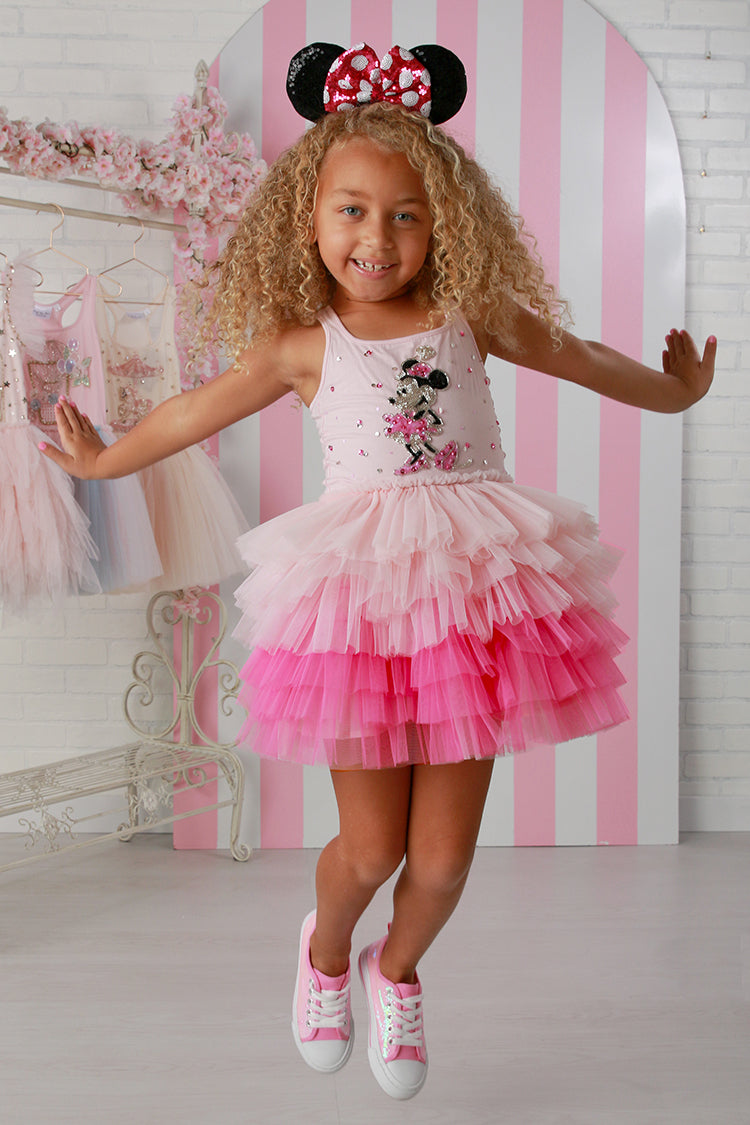 Young girl in a pink and white dress with Minnie Mouse design, standing in front of a pink and white striped wall.