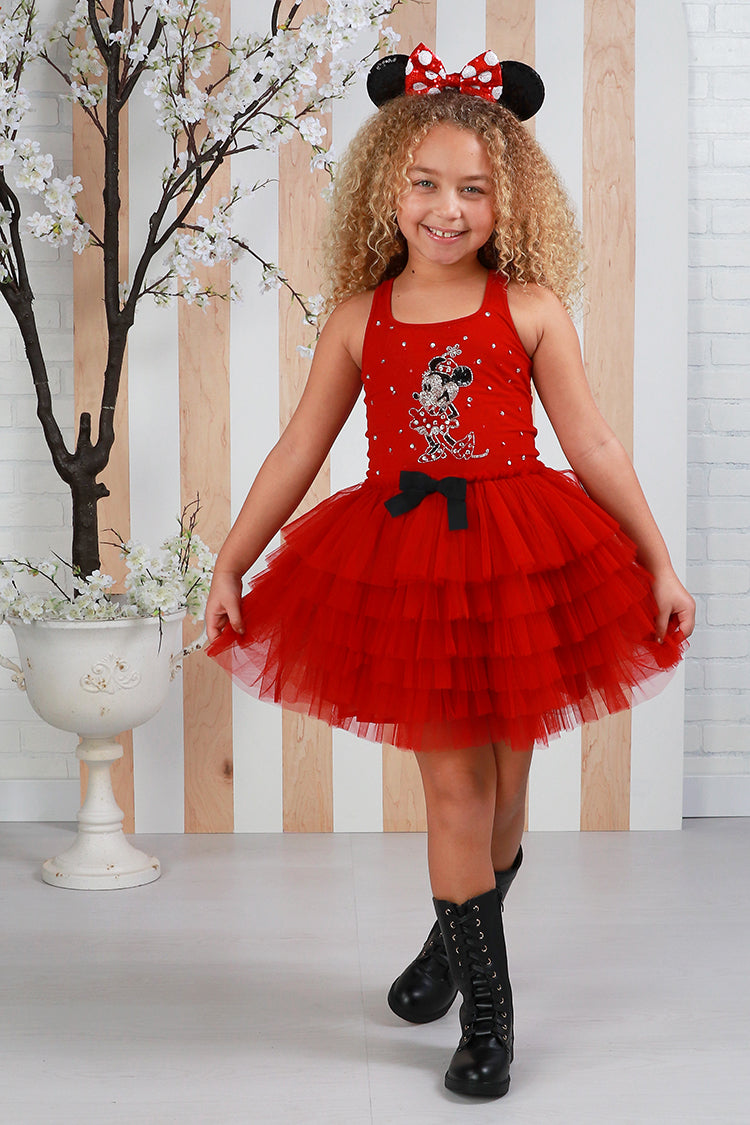 Young girl wearing a red dress with black polka dot headband, standing against a wooden paneled wall.