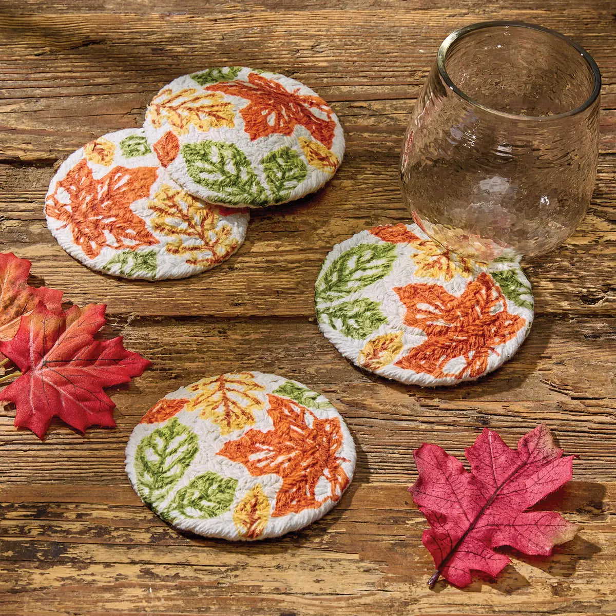 Set of four leaf-patterned coasters on a wooden surface with a glass and additional leaves.