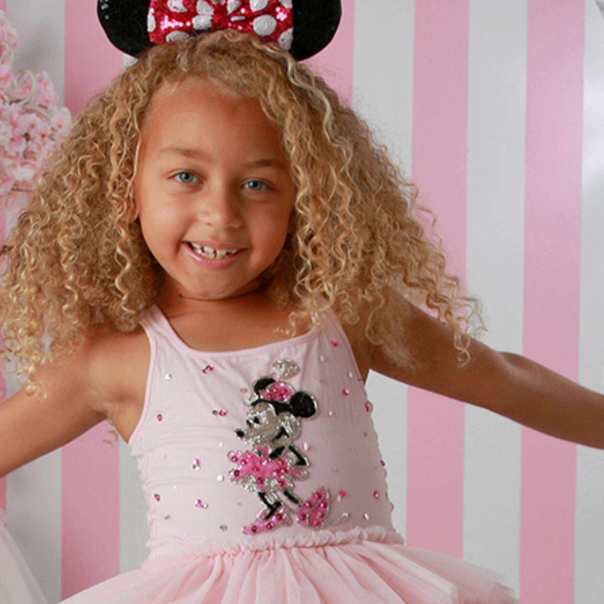 Young girl wearing a pink dress with a Minnie Mouse design and Minnie Mouse ears against a pink and white striped background.