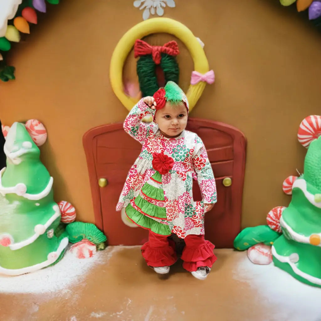 Child in festive outfit standing in front of a gingerbread house with candy decorations.