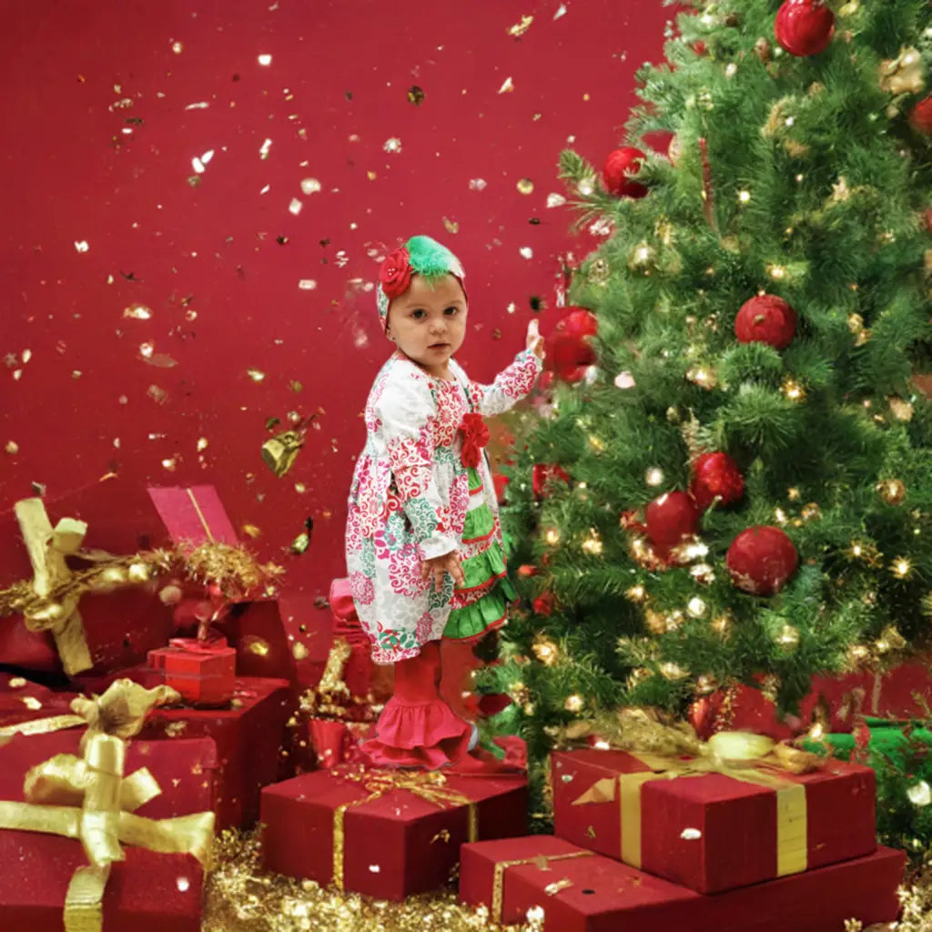 Child in festive outfit standing next to a decorated Christmas tree with red gifts on a red background