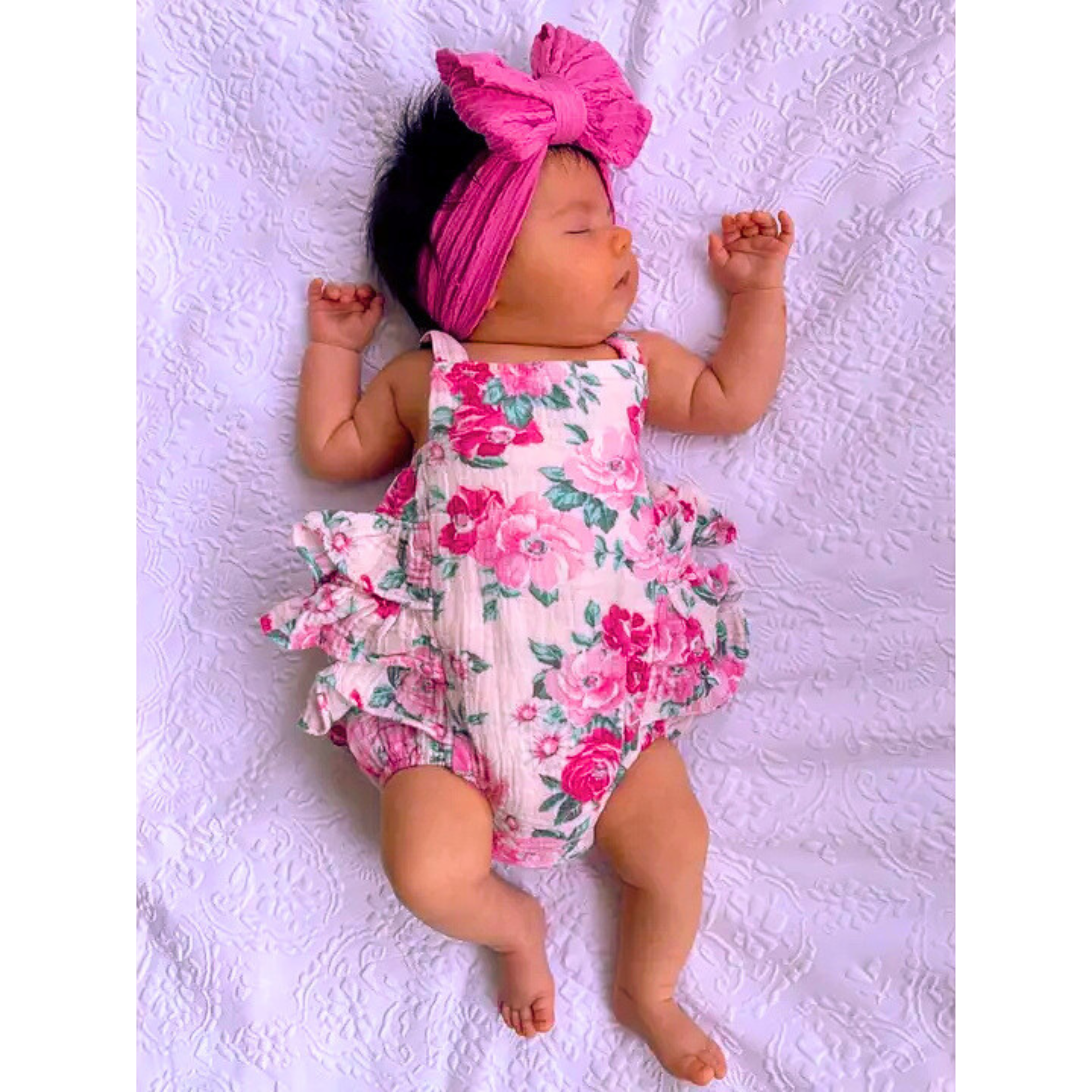 Newborn baby wearing a floral romper and pink headband on a white textured surface