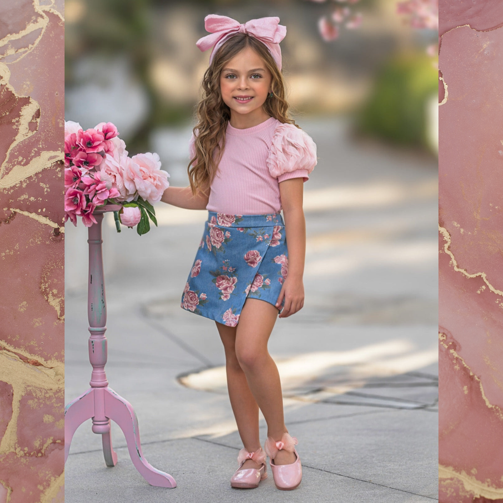 Girl with pink bow wearing a pink puff sleeve top, rose print skort, and pink shoes on a pink background