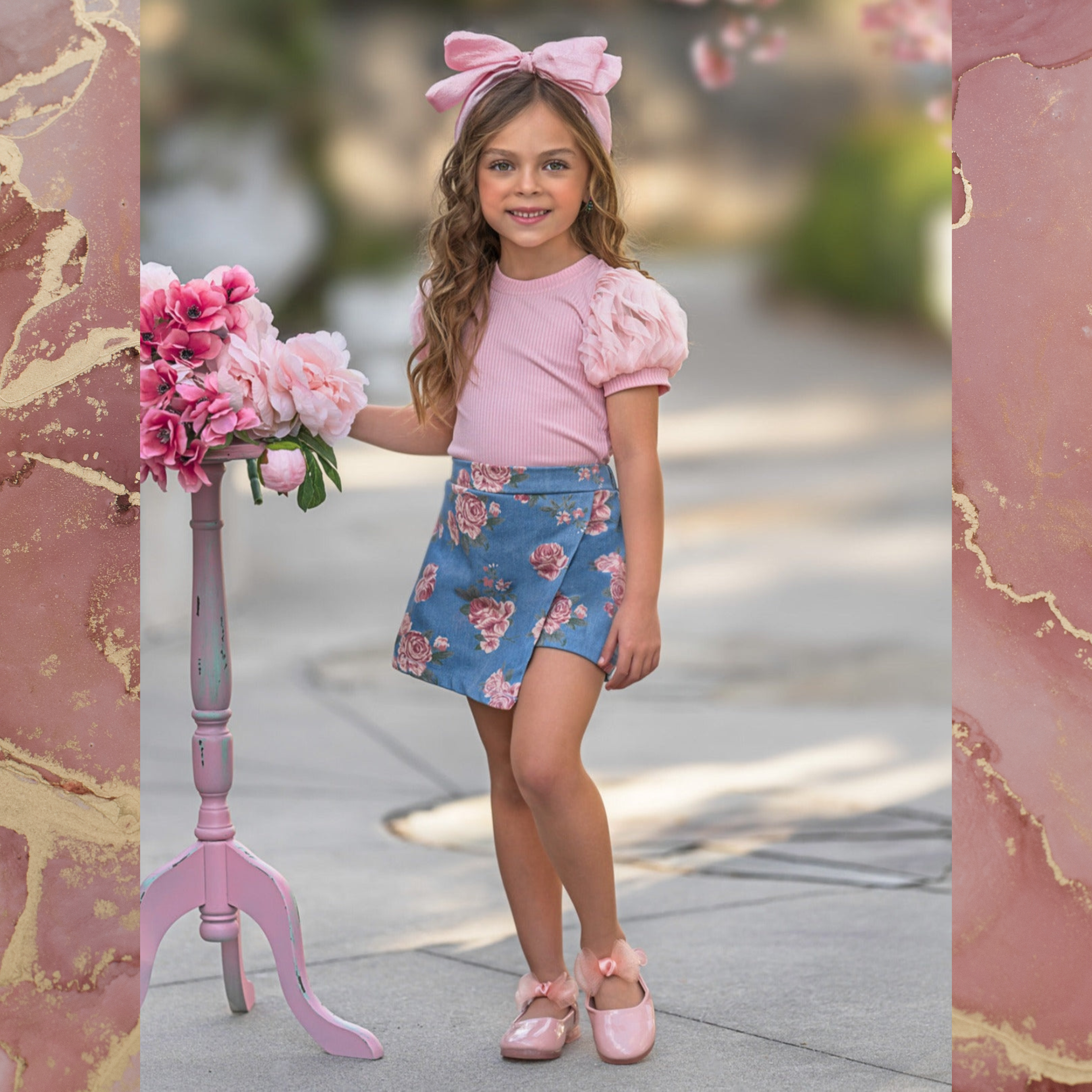 Girl with pink bow wearing a pink puff sleeve top, rose print skort, and pink shoes on a pink background