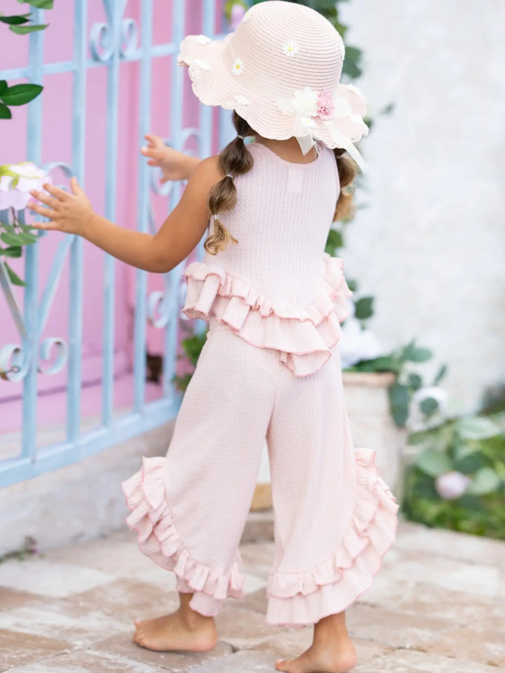Young girl in a pink outfit with ruffles and a matching hat, standing in front of a pink gate.