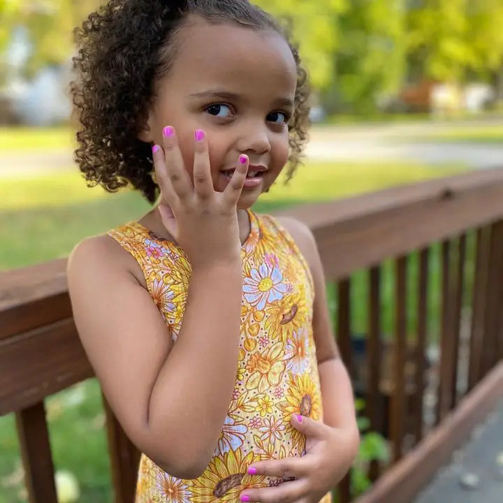 Young girl in a yellow floral dress standing on a wooden deck with greenery in the background