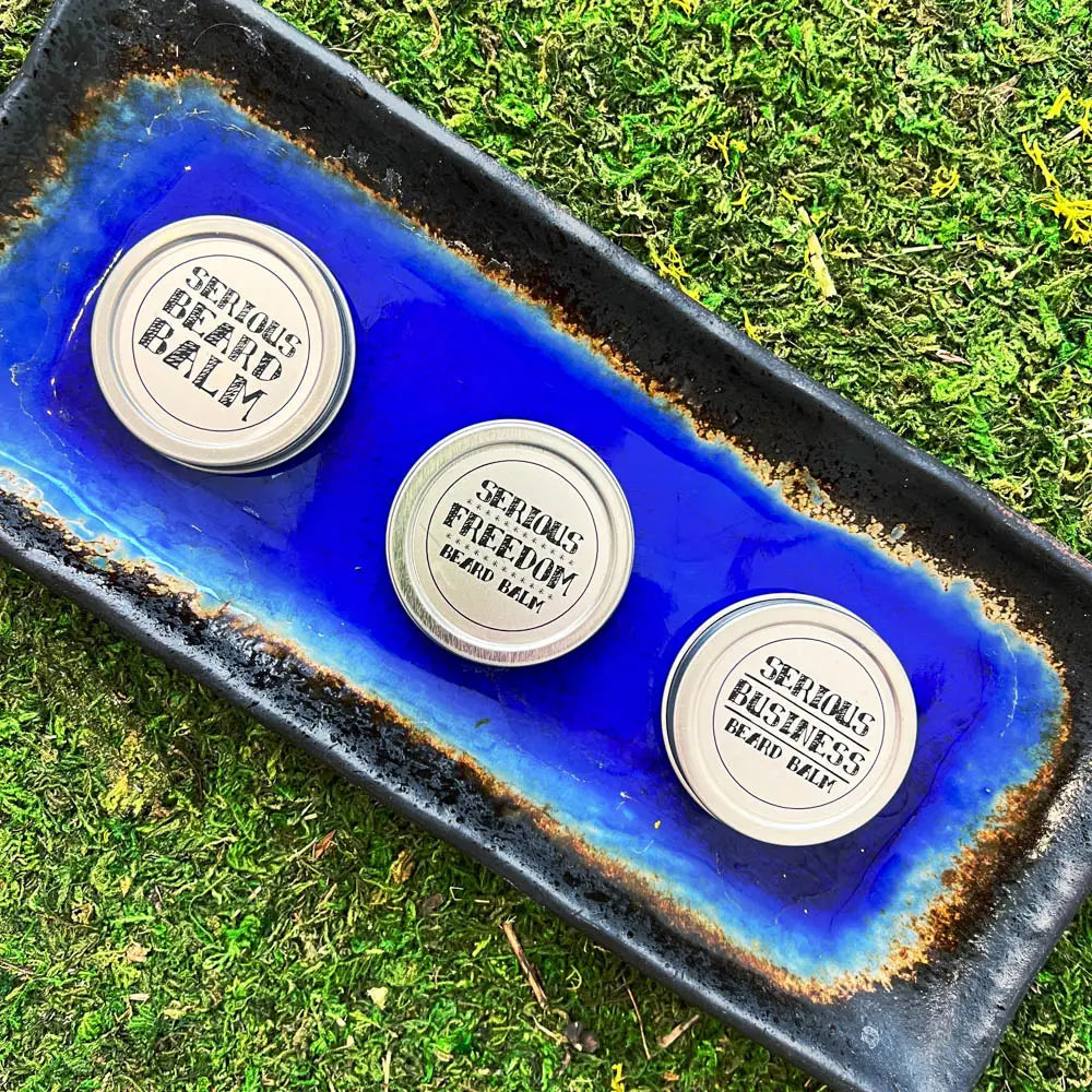 Three containers of 'Serious Freedom' balm on a blue and black tray with grass in the background.