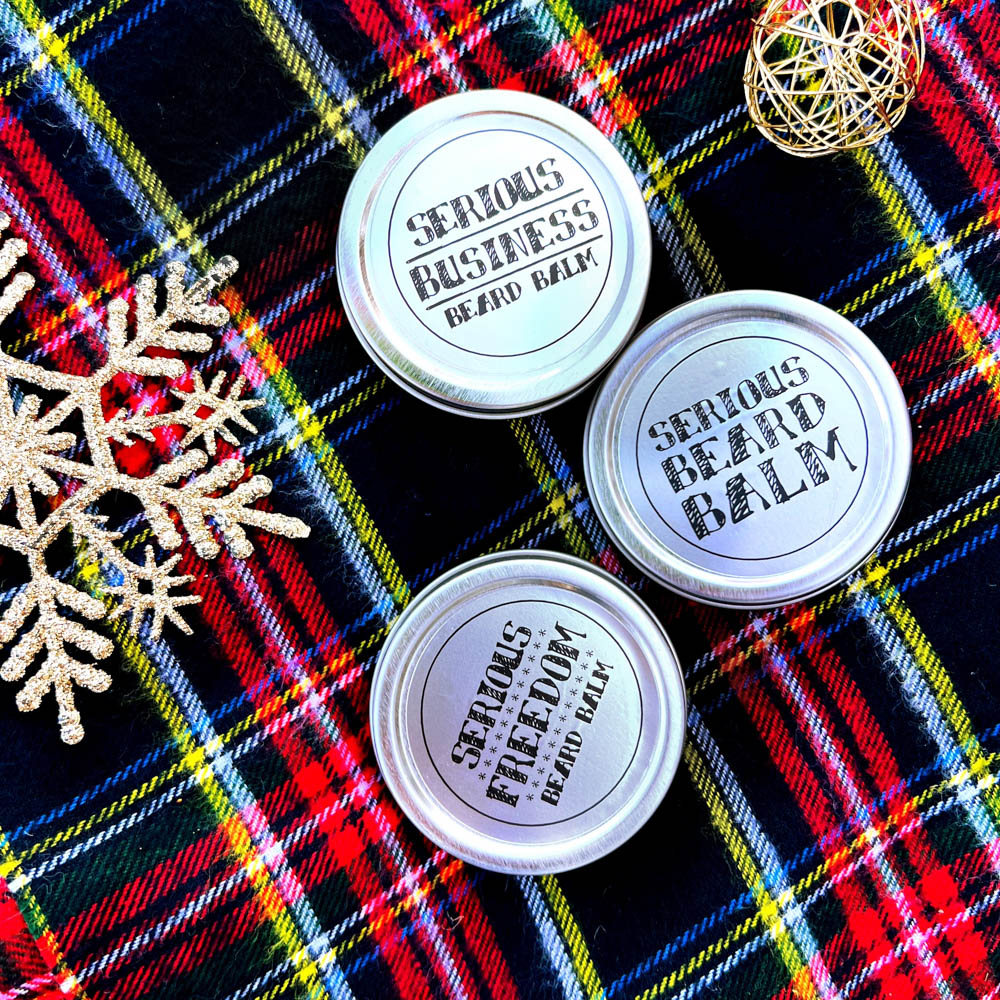 Three tins of 'Serious Beard Balm' on a plaid fabric background with decorative elements.