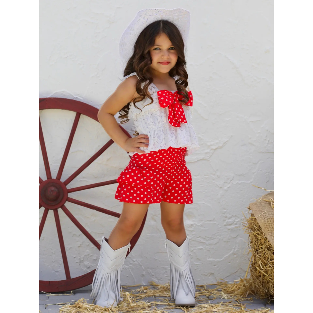 Young girl in a cowboy outfit with a white top and red polka dot shorts, standing in front of a white wall with a red wagon wheel.