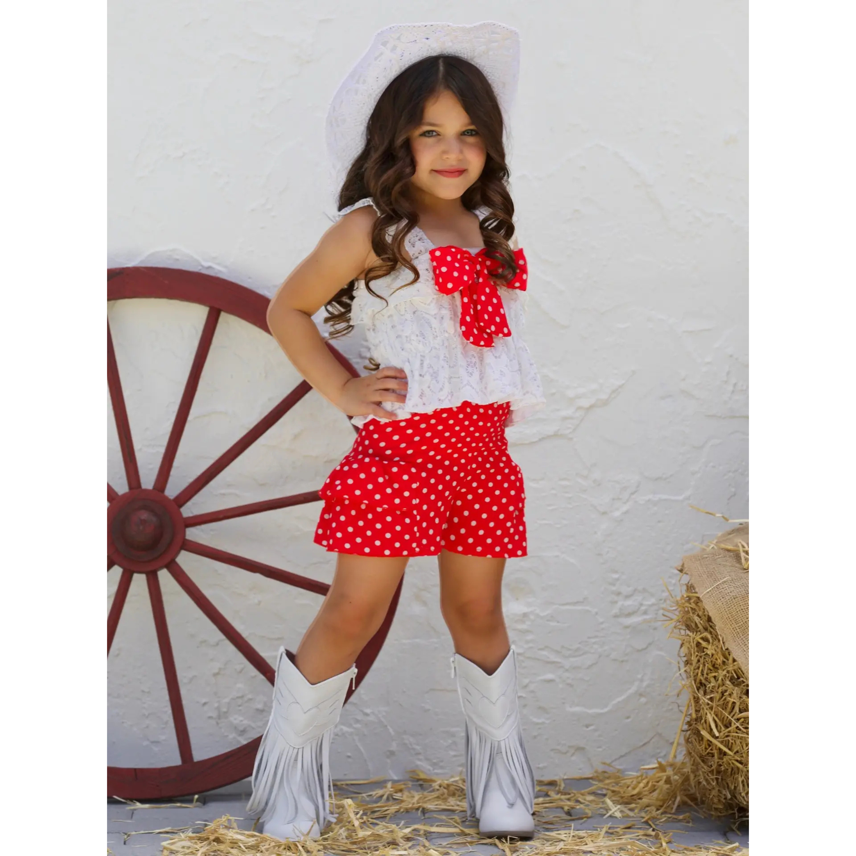 Young girl in a cowboy outfit with a white top and red polka dot shorts, standing in front of a white wall with a red wagon wheel.