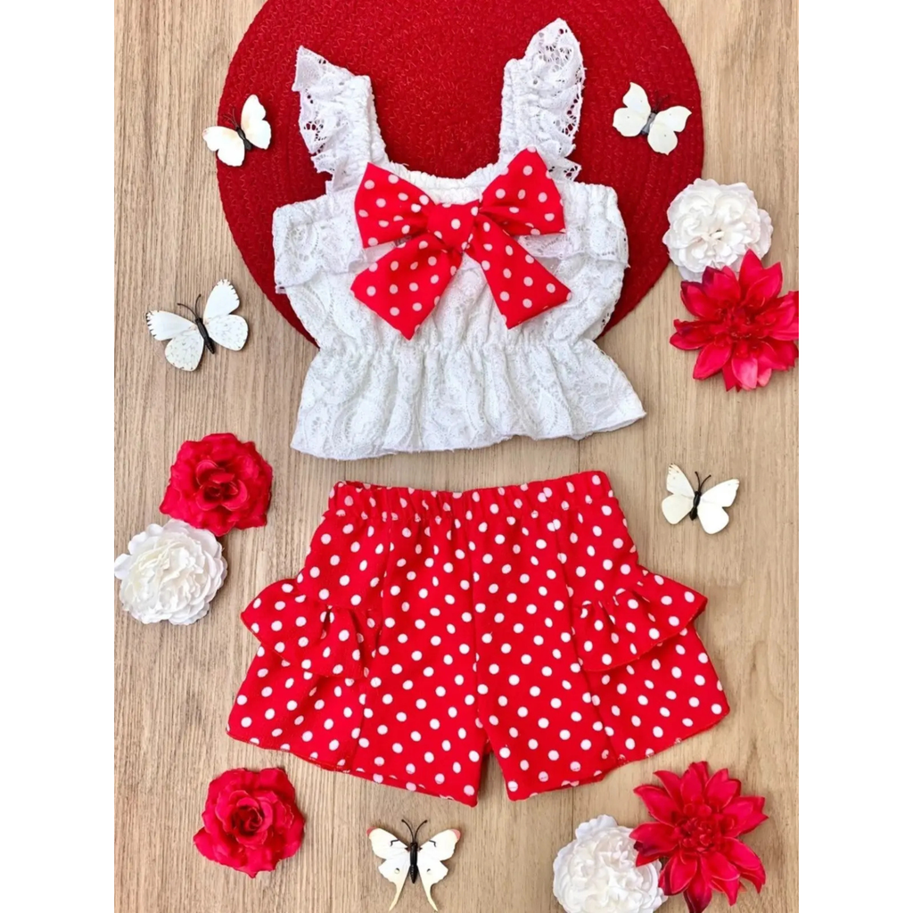Children's outfit with white top and red polka dot shorts on a wooden surface with flowers and butterflies.