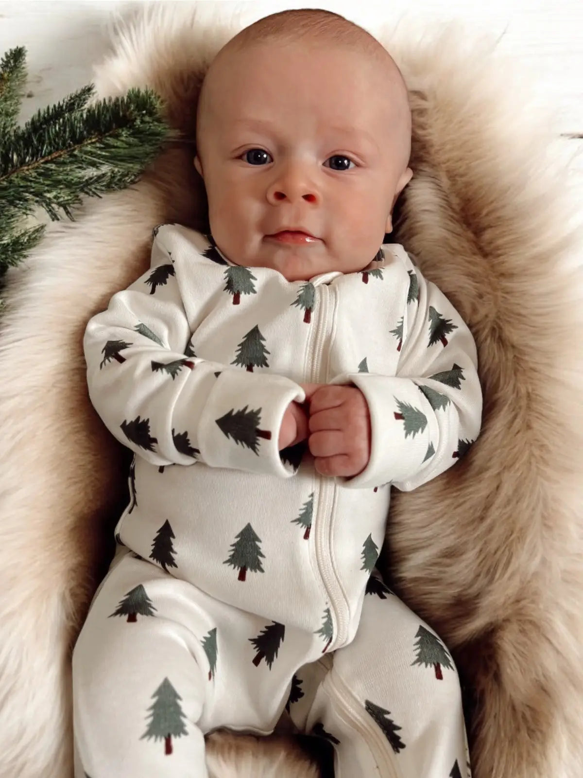 Baby wearing a white onesie with tree patterns, lying on a fluffy beige surface.