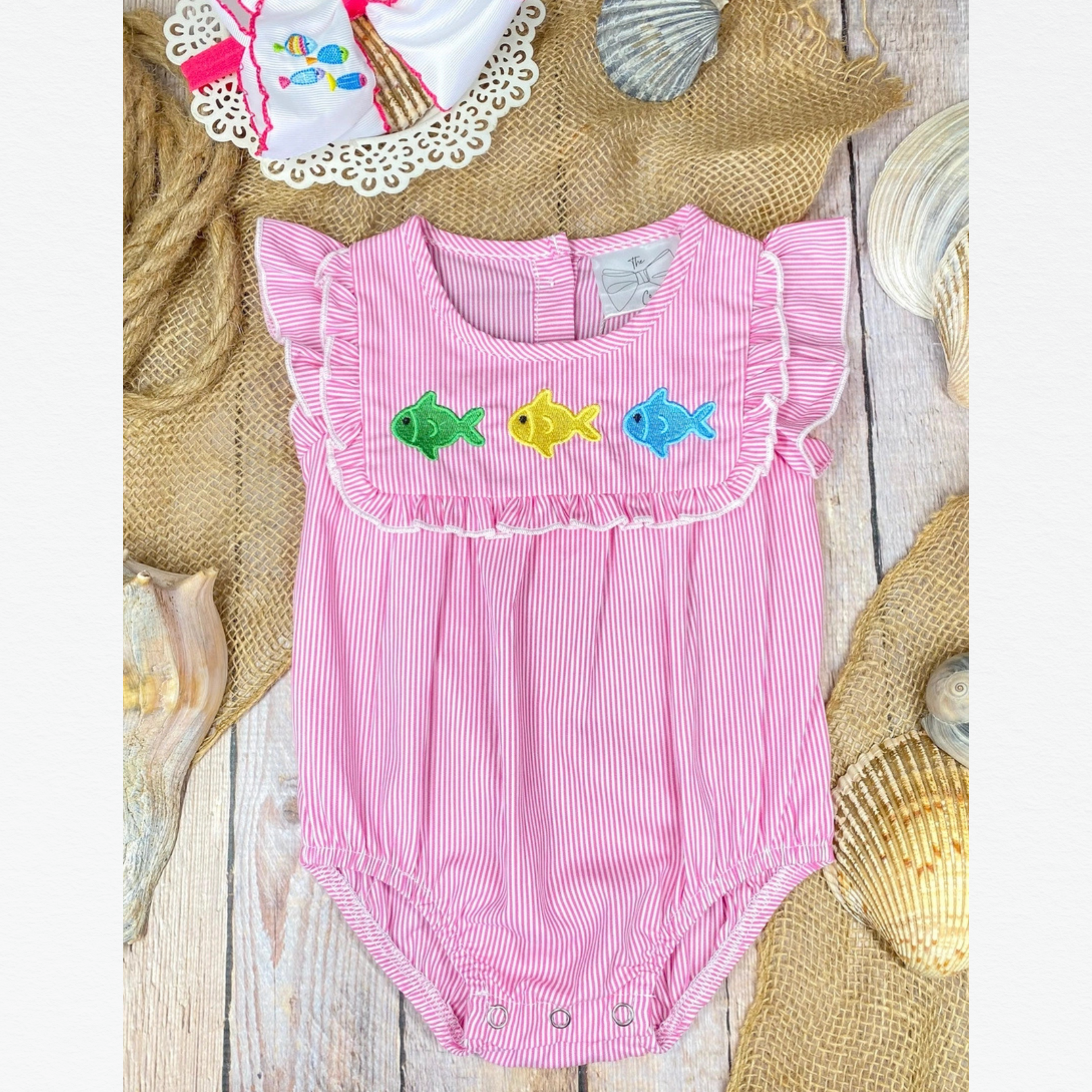 Pink baby romper with fish embroidery on a wooden surface with seashells.