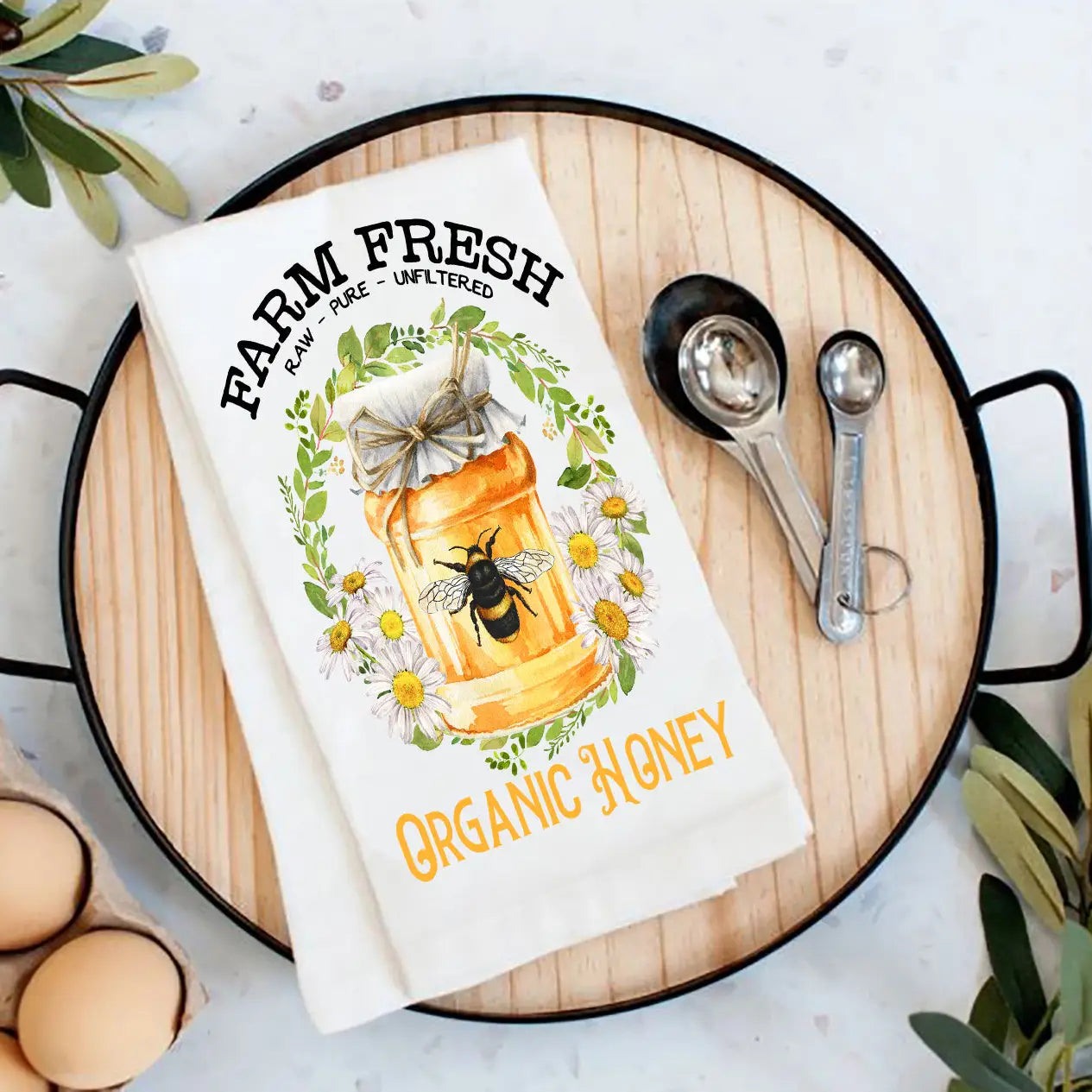 Tea towel with honey jar design and text on a wooden board with measuring spoons and eggs.
