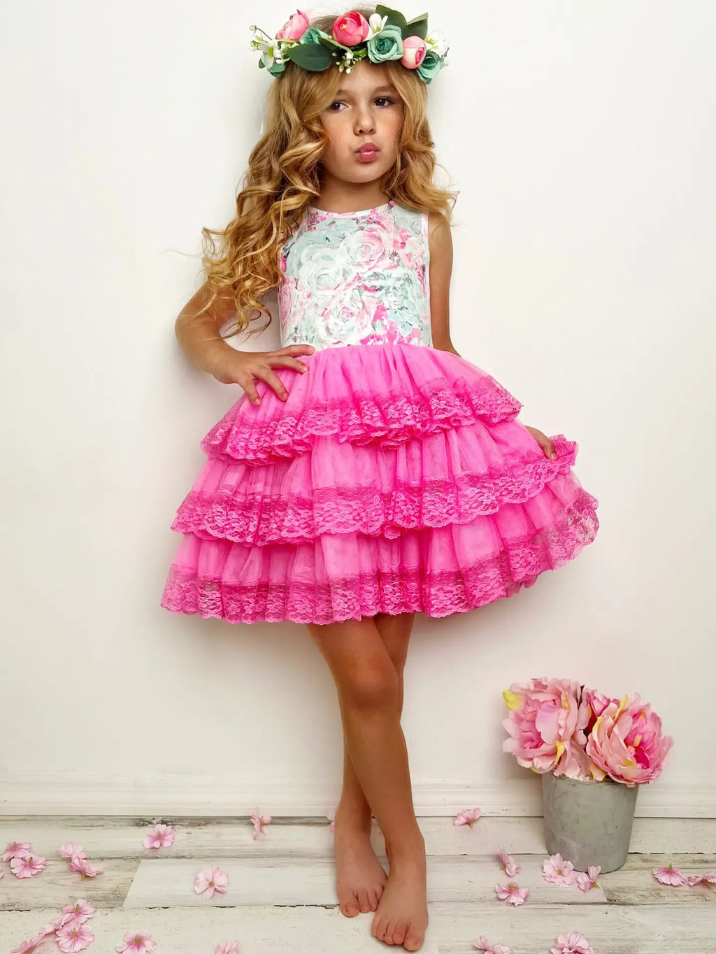 Young girl wearing a floral top and pink ruffled skirt with a flower crown, standing on a wooden floor with flowers.