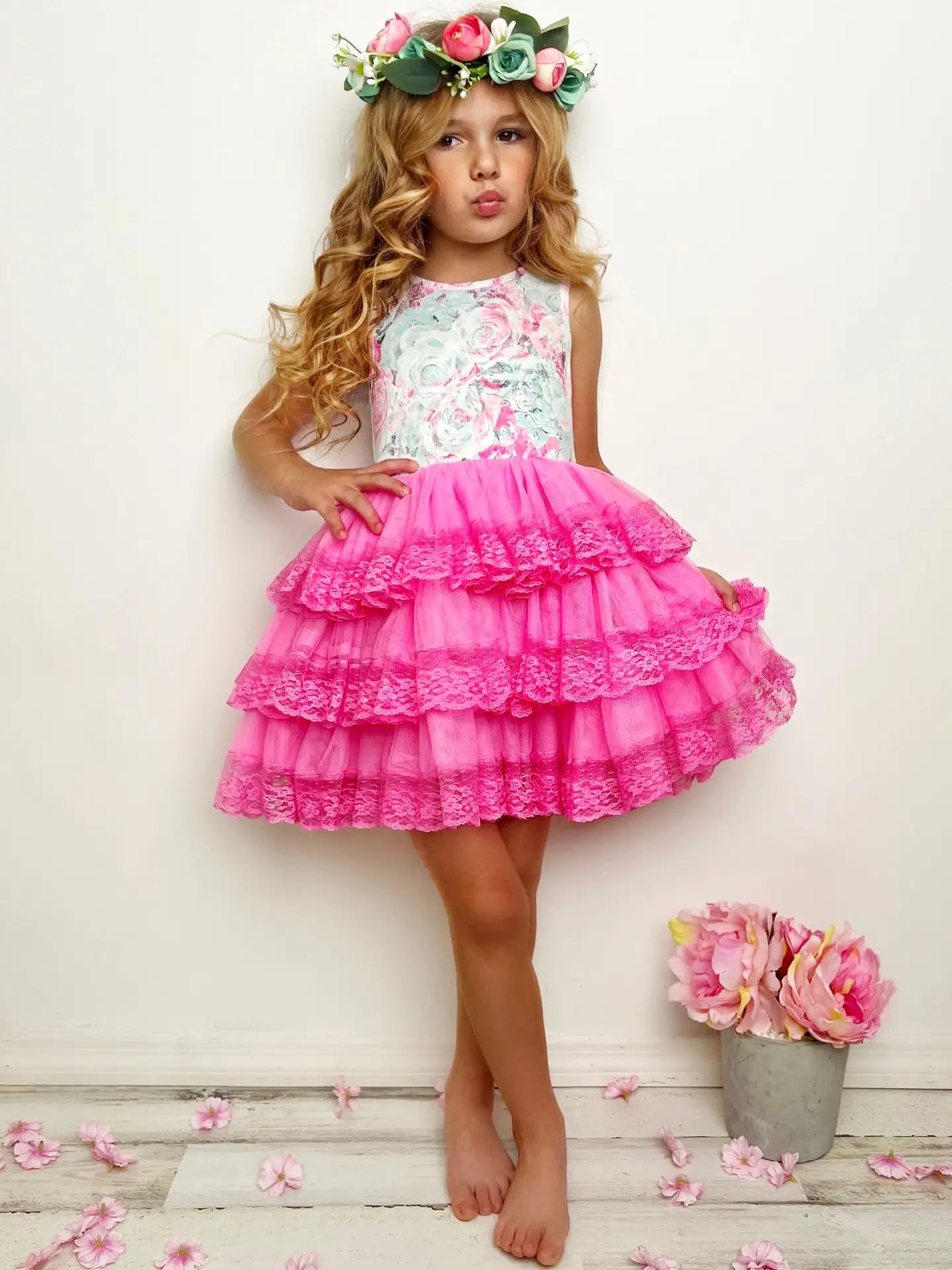Young girl wearing a floral top and pink ruffled skirt with a flower crown, standing on a wooden floor with flowers.