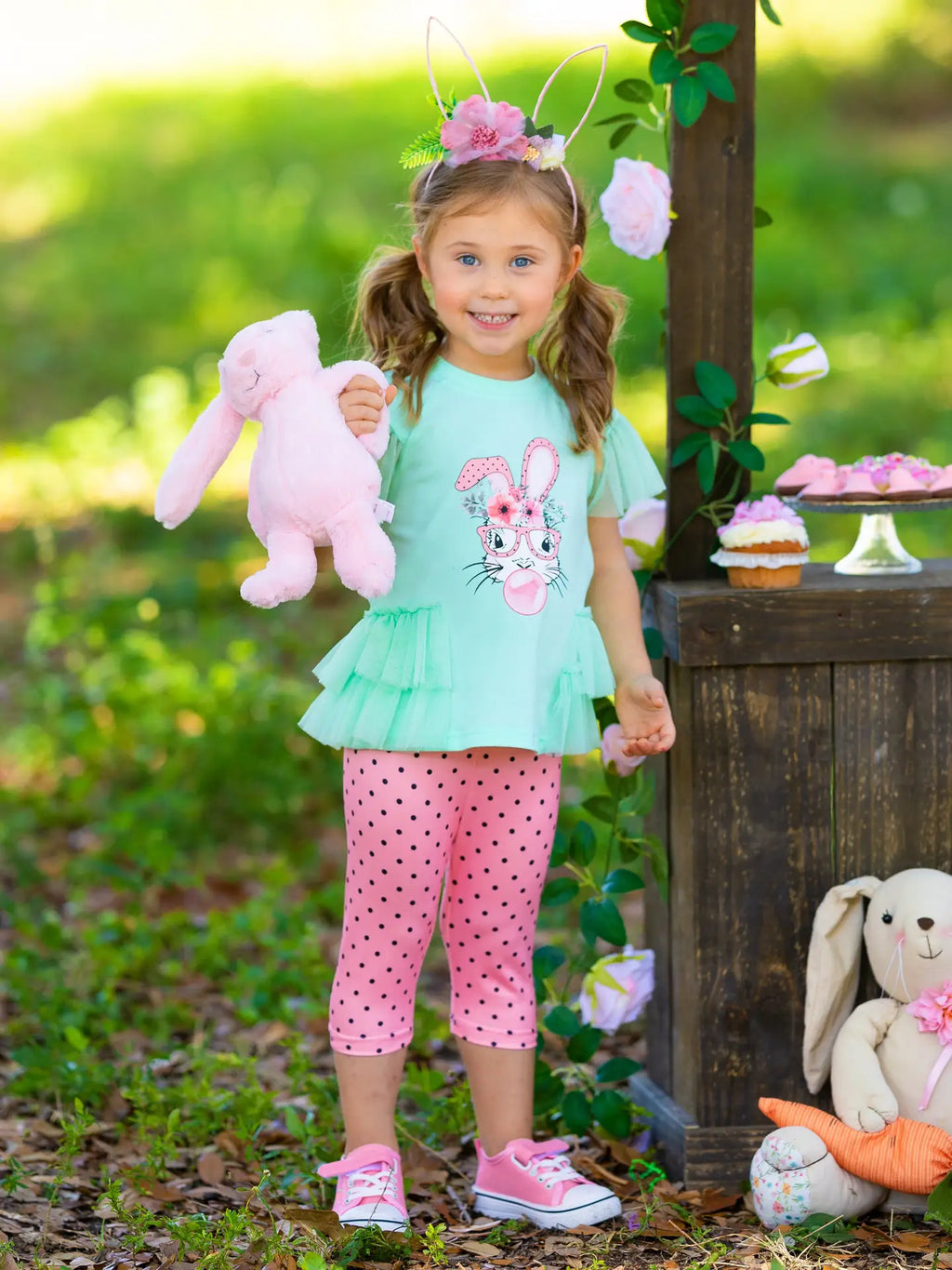 Young girl in a light blue top and pink polka dot pants holding a pink bunny toy outdoors.
