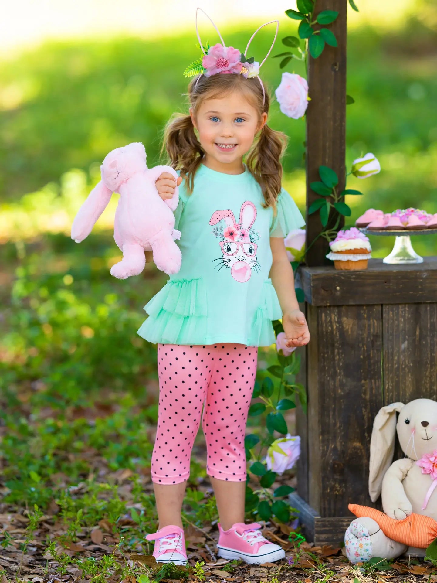Young girl in a light blue top and pink polka dot pants holding a pink bunny toy outdoors.