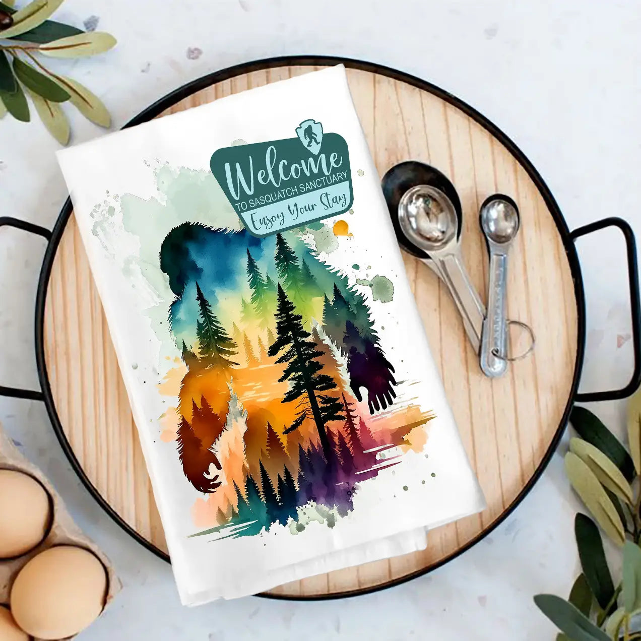 Colorful towel with big foot and forest design on a wooden cutting board with measuring spoons.