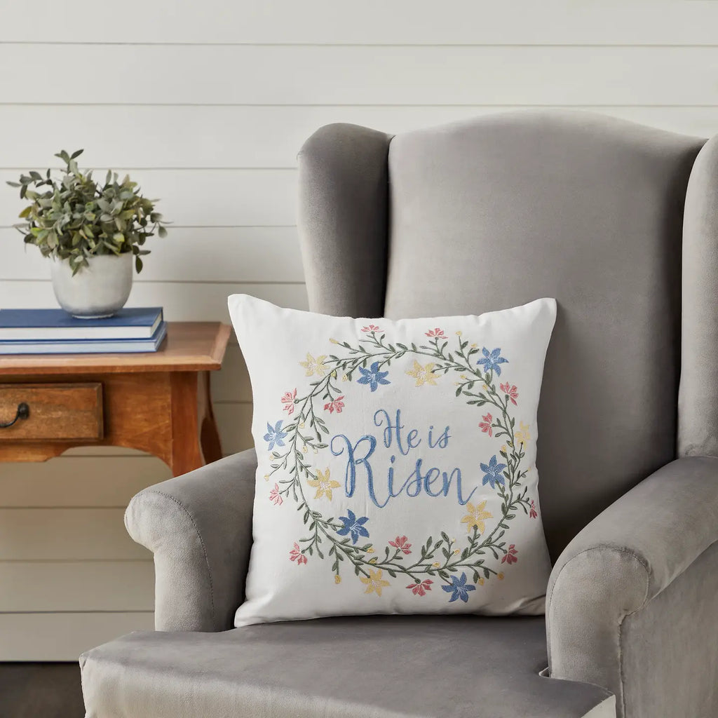 Decorative pillow with 'He is Risen' text on a gray armchair in a home setting.