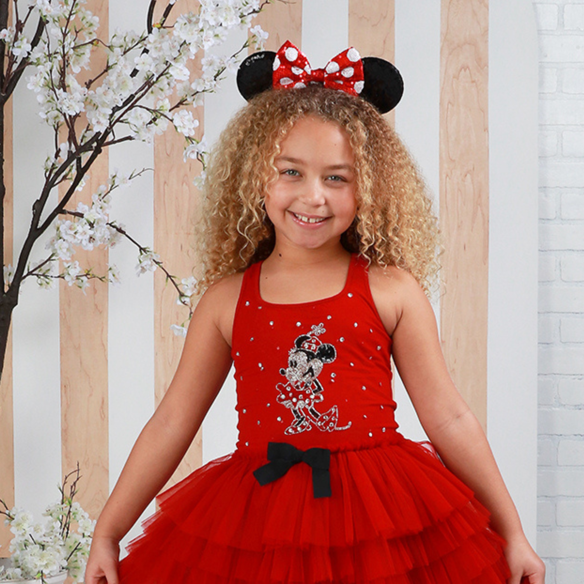 Young girl wearing a red dress with Minnie Mouse design and black bow, standing against a light wooden panel background.