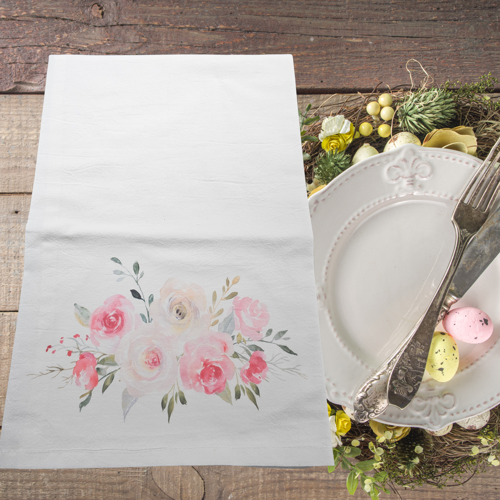 Floral flour sack dish towel on a wooden table with cutlery and decorative elements.
