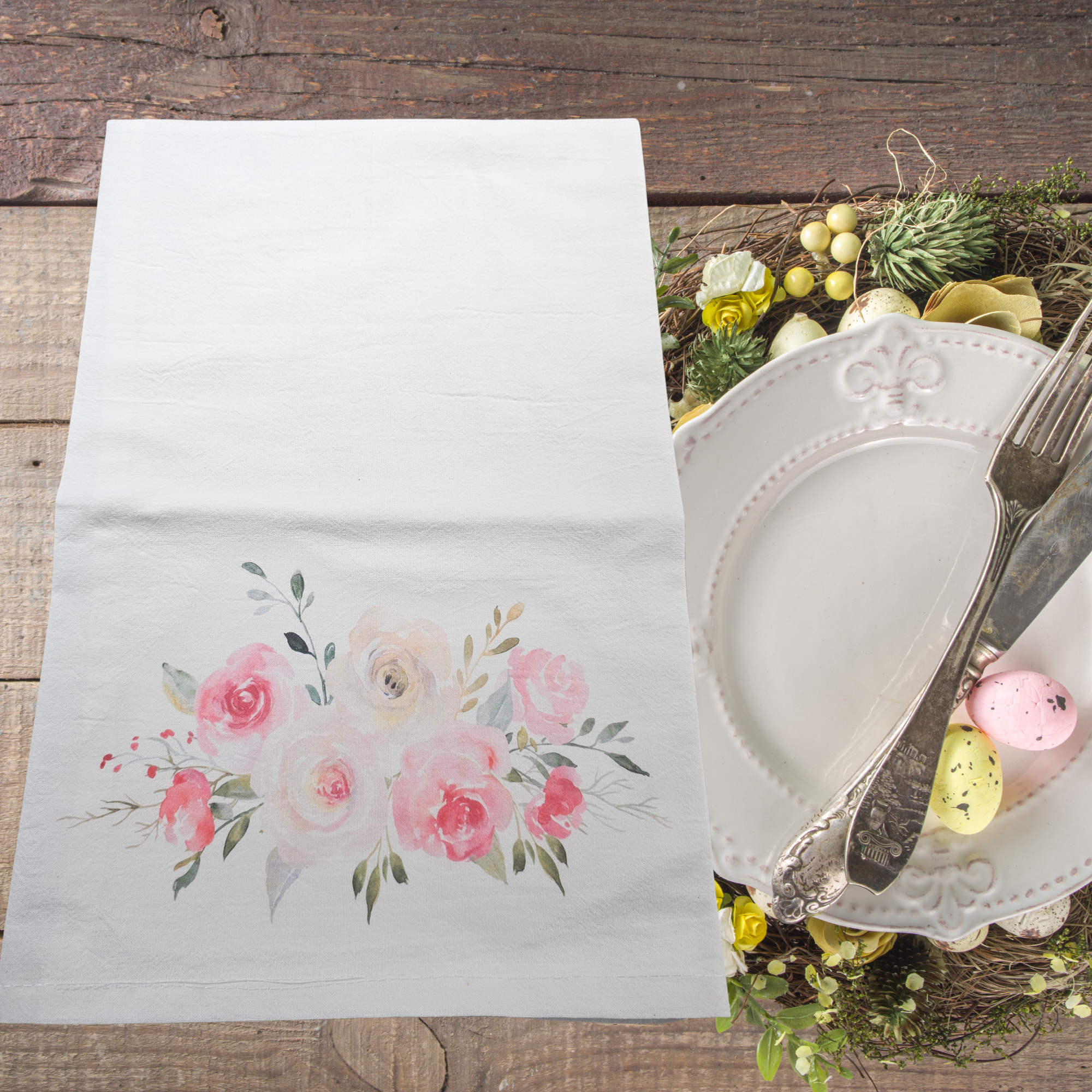 Floral flour sack dish towel on a wooden table with cutlery and decorative elements.
