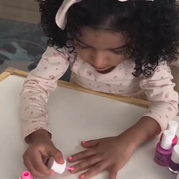 Child applying nail polish to a small object with various nail polish bottles around.