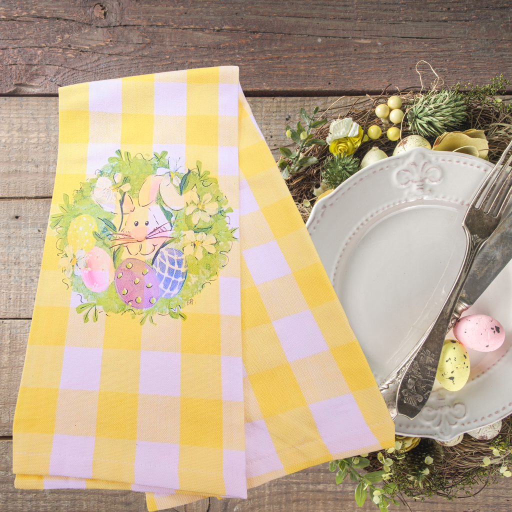 Yellow and white checkered dish towel with Easter design on a wooden table with cutlery and eggs.